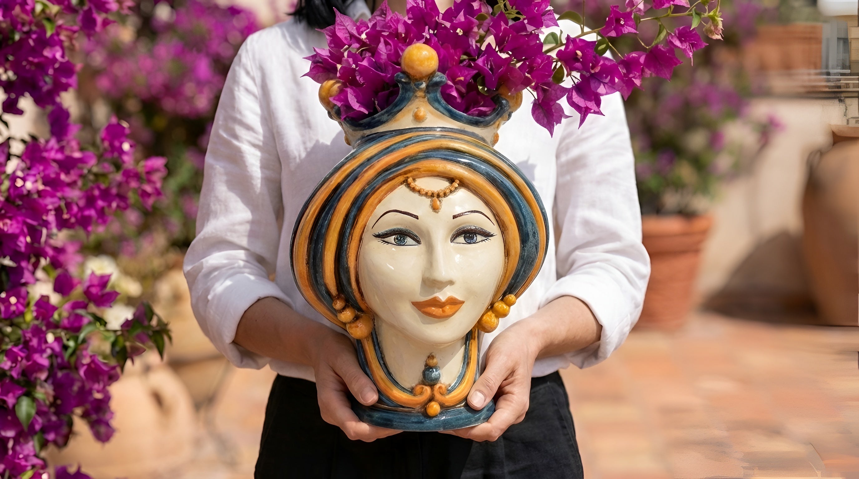 Testa di moro femminile con corona, turbante blu e arancio, tenuta fra le mani davanti a una bougainvillea in fiore