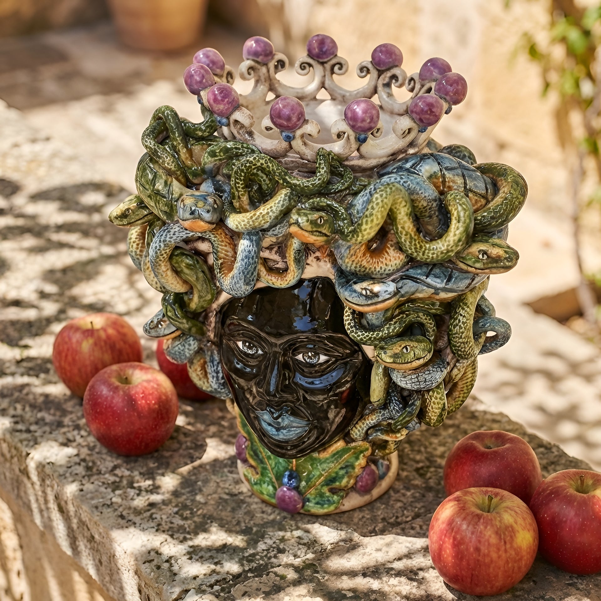 Handmade Teste di Moro vase with snakes and crown, traditional Sicilian ceramic art, displayed with apples on stone.