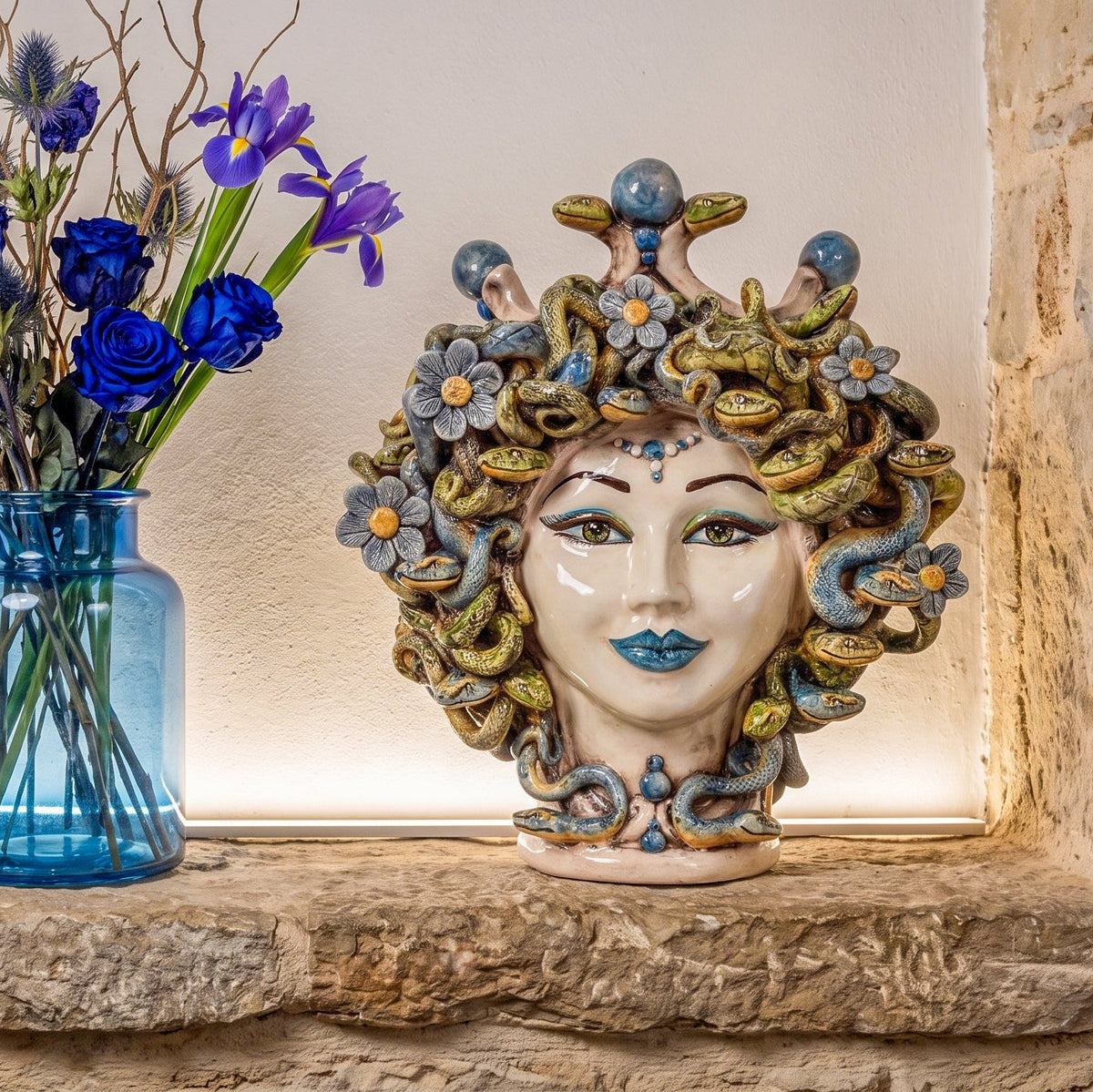 Medusenkopf mit Blumen auf einer Steinablage neben einer Vase mit blauen Iris