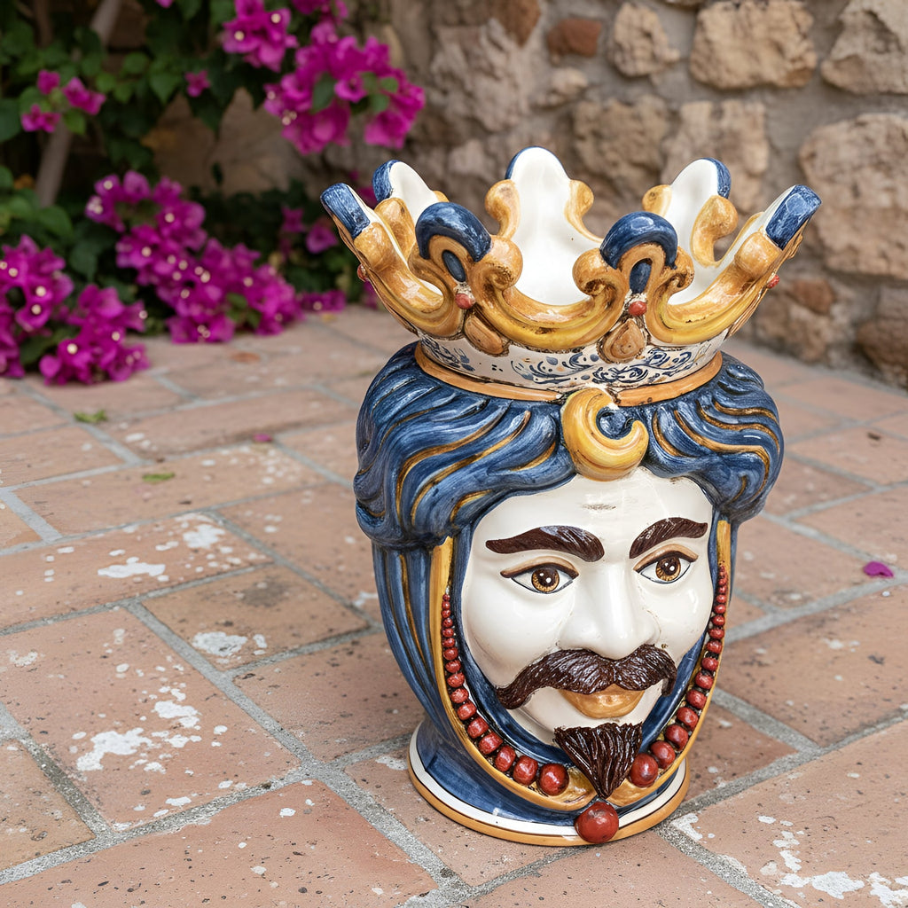 Handcrafted Teste di Moro ceramic vase with crown, blue hair, and red beads, set against a stone wall and flowers.