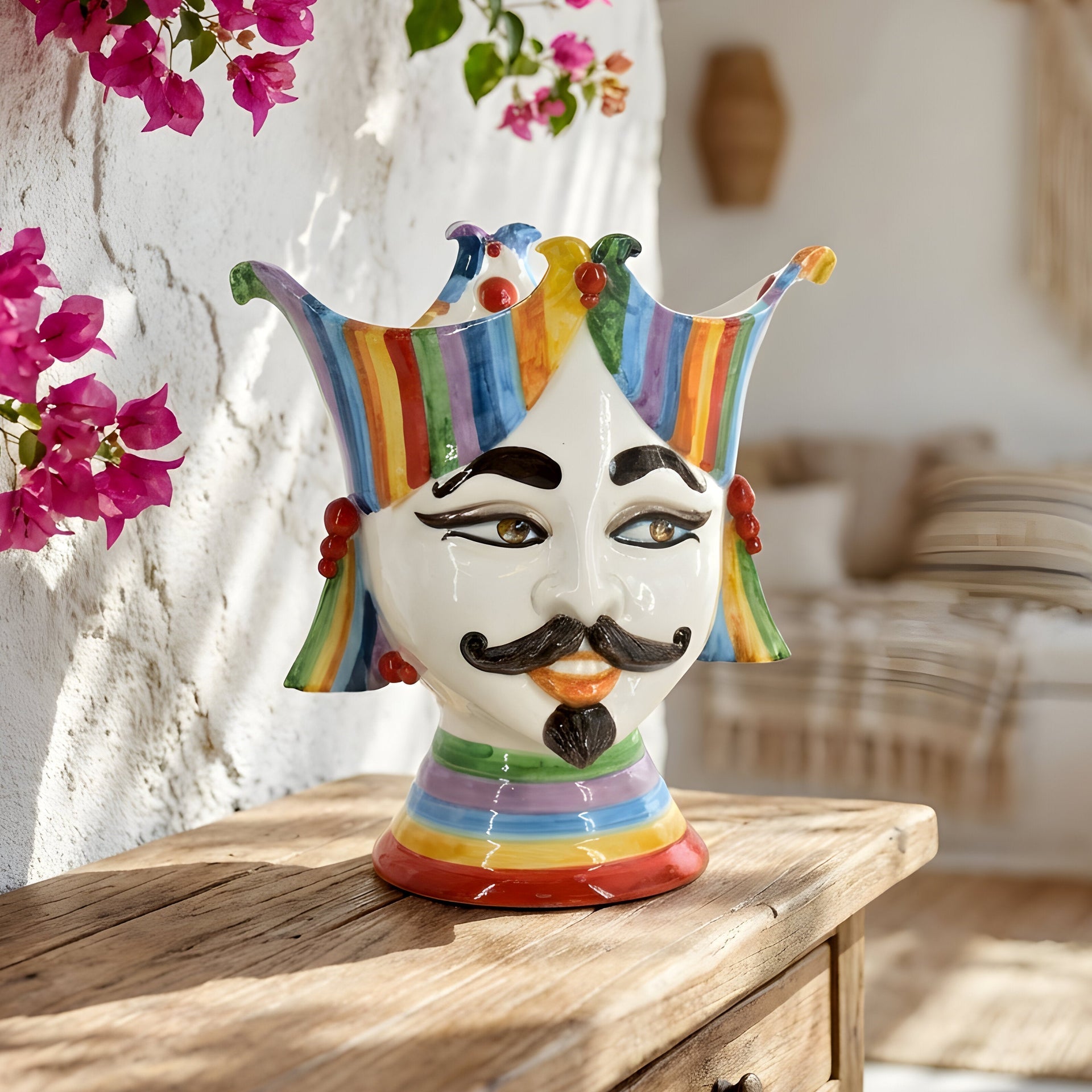 Handmade ceramic Testa di Moro vase with colorful jester crown, displayed on rustic wood table with pink flowers.