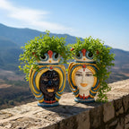 Handmade Teste di Moro planters with vibrant faces, lush greenery, set against scenic hills—perfect Sicilian decor.