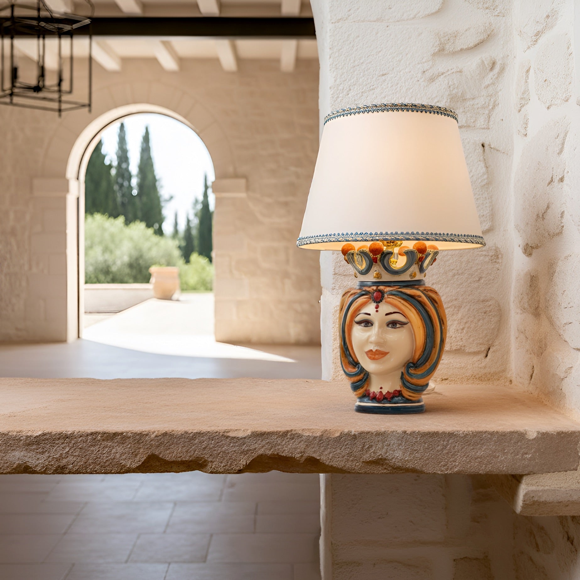 Handmade Sicilian Testa di Moro table lamp with colorful ceramic face, crown, and cream shade on stone ledge